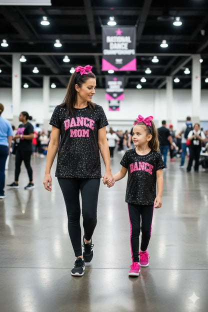 Woman and young girl holding hands, both wearing matching 'DANCE MOM' and 'DANCE' shirts in a large indoor venue.