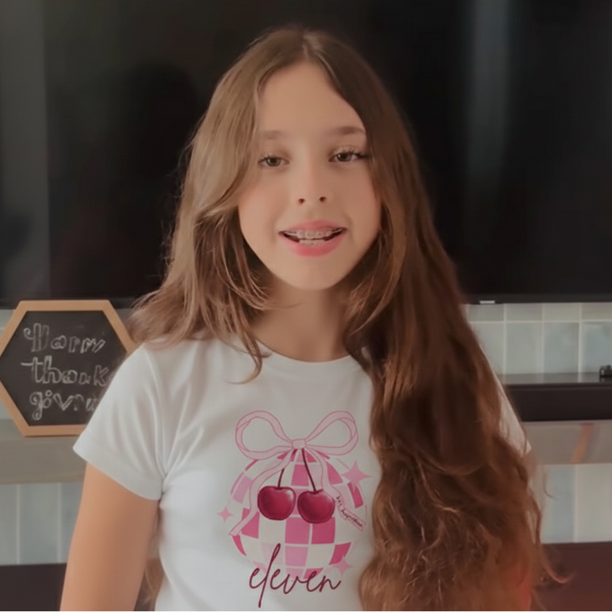 Young girl wearing a white t-shirt with a graphic design of cherries and the number 'eleven'.